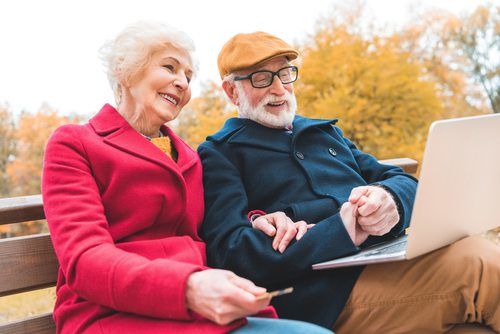 smiling senior man and woman sitting on a bench outside and using a laptop
