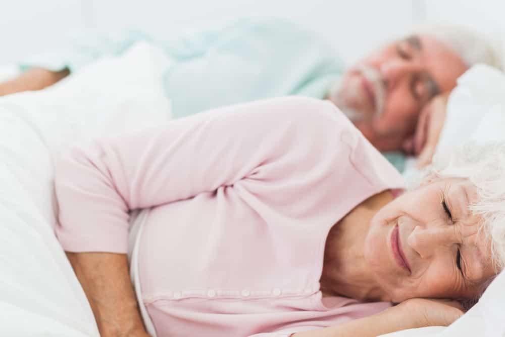 Senior-Woman-Sleeping-on-Her-Side-Softly-Smiling-Husband-Asleep-Behind-Her