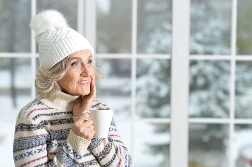 Smiling-senior-woman-wearing-knit-hat-holding-mug-in-front-of-window-revealing-snowy-scene