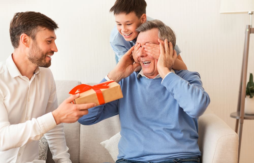 Man giving gift to senior man, whose eyes are covered by smiling boy