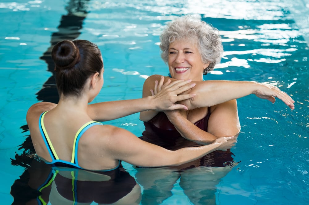 Senior woman stretching in pool with help of instructor